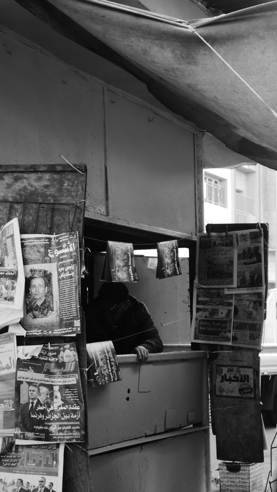 Kiosque à journaux, présentoirs couverts de titres, le vendeur en retrait