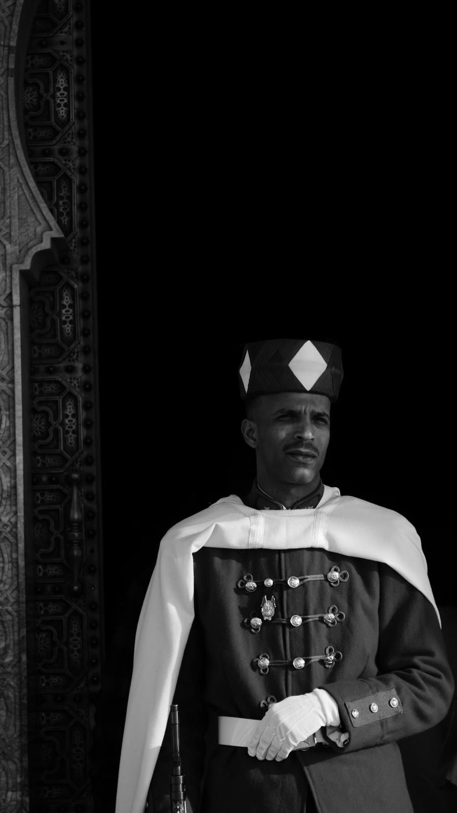 Garde en tenue d'apparat, cape claire et coiffe, devant une embrasure sombre